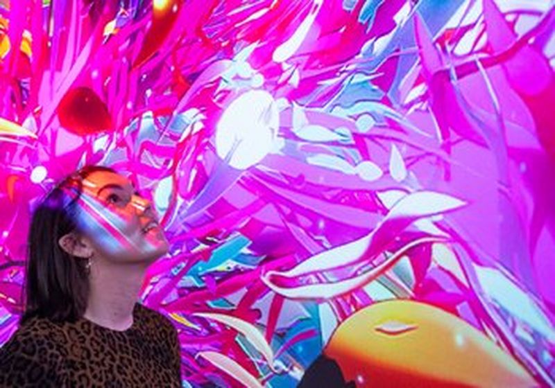 A woman’s head and shoulders as she stands in the middle of a brightly coloured projection of leaves and shapes in pink, red, purple and blue.