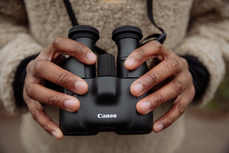 A pair of hands hold out a set of black Canon 8x20 IS binoculars in front of their body.