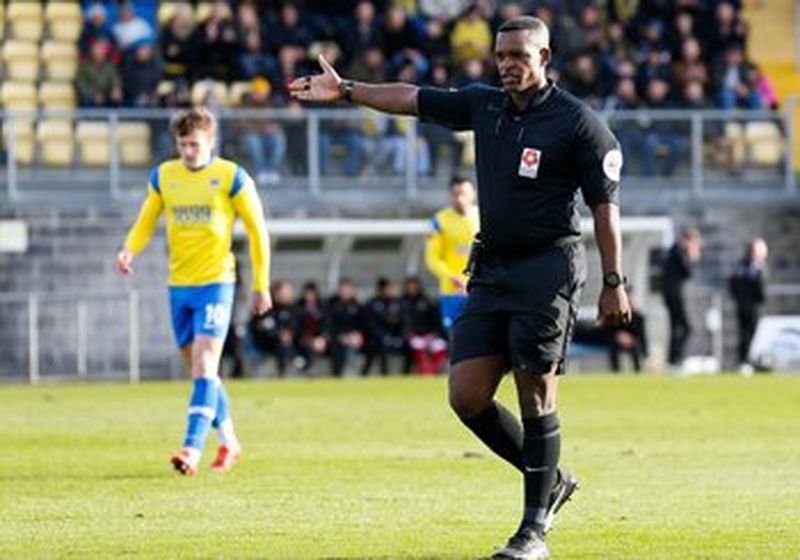 A man in a black referee’s uniform stands on a football pitch with his right arm outstretched. To his left and rear is a footballer wearing a yellow top, blue shorts and matching socks. There are blurred figures in the background who appear to be sat in the players dugout. Behind them are the crowd, who are watching the match.