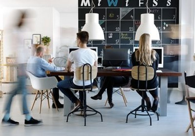 A modern office environment, with a chalkboard wall as a team calendar, plants and natural light. Employees sit around a dining style table, wearing jeans and t-shirts.