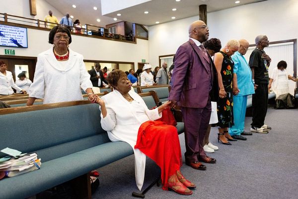 Congregants stand holding hands at church – one woman sits with a red shawl across her knees. Photo by Laura Morton.