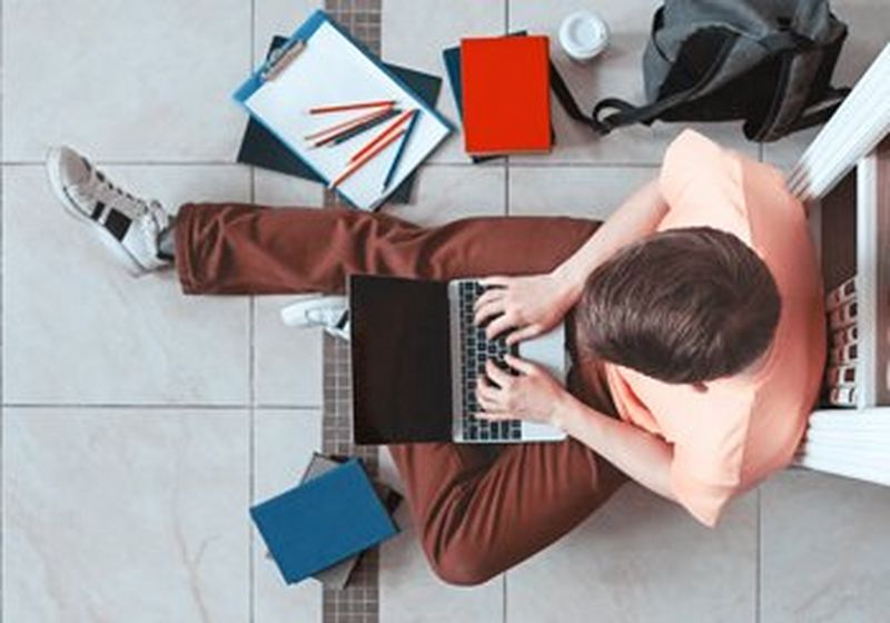 Taken from above, a teenage boy wearing a peach t-shirt and brown trousers sits on a tiled floor using a laptop. He has shelves behind him and stationery around him on the floor.