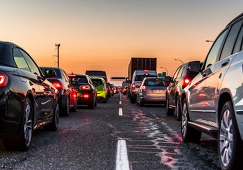 A photo taken from the centre of a traffic jam, standing on the central white live, viewing cars on left and right from behind. It is sunset and both cars and lorries are at a standstill, reflecting each other and their lights in their paintwork. In the distance there are power lines connected to telegraph poles.  