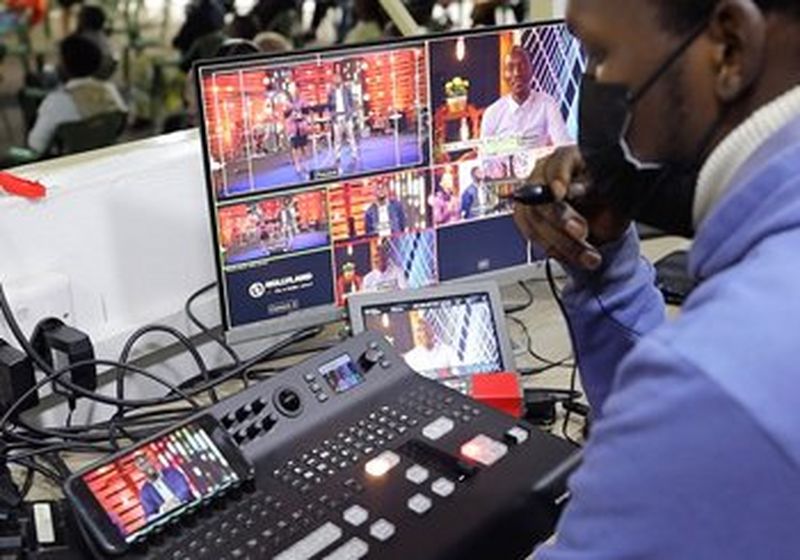 On the right, the side of a man wearing a black medical-style mask and a blue top is pictured. He is overseeing an audio-visual control panel of buttons, some of which are lit. There is also a monitor showing what is being filmed and cables feeding into plug sockets.