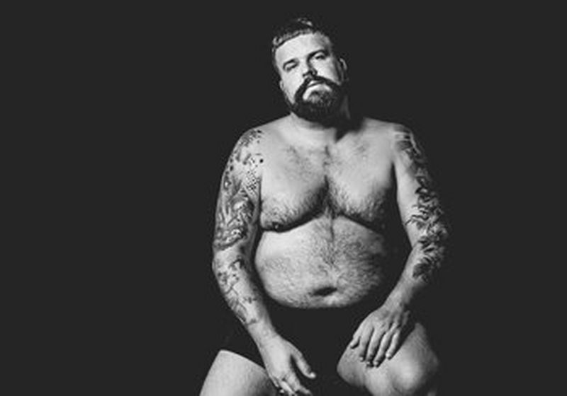 A bearded man sits against a black background. He is wearing just black shorts and has tattoos on both arms.