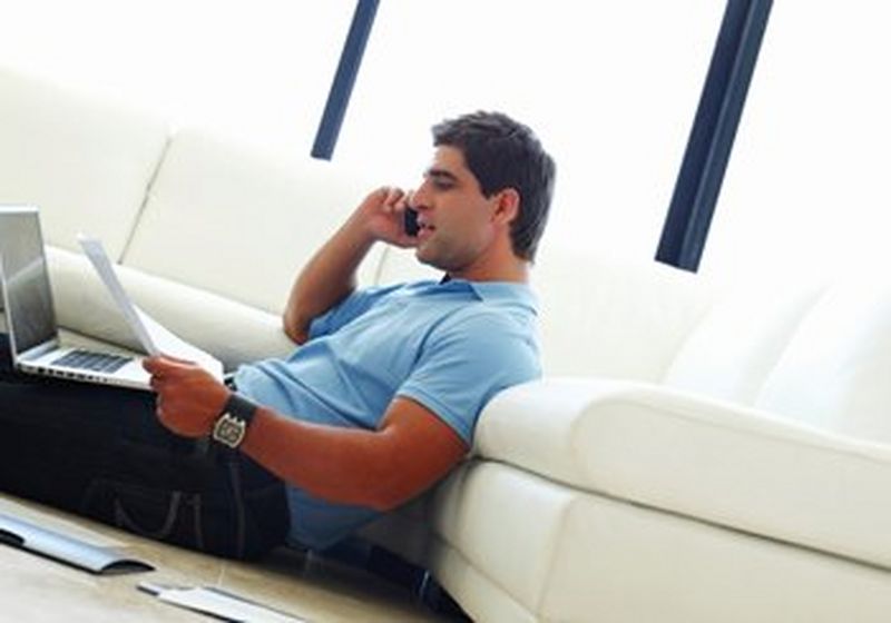 A man, clearly working from home with a laptop on his lap, speaking on a mobile phone.