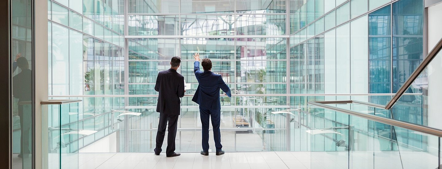 Two suited men with their backs to the camera gaze out through a large glass office building, with one pointing upwards.