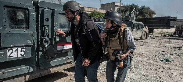 Photojournalist Veronique de Viguerie wears a flak jacket and helmet, her Canon camera flung around her neck, and ducks beside a military vehicle. She is with a man in plain clothes who is also wearing a helmet.