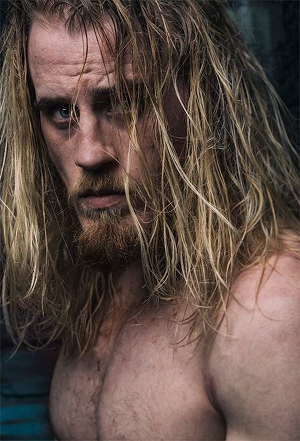 Wrestler Mark Haskins stands by a wooden door, long hair wet with sweat, and looks at the camera.