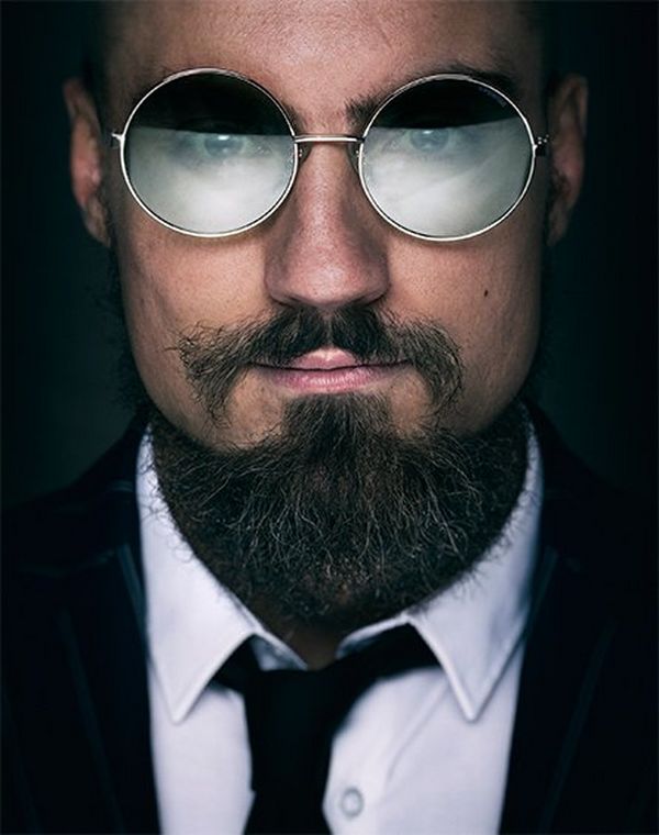 A close-up portrait of wrestler Marty Scurll wearing a suit, tie and round sunglasses, looking at the camera.