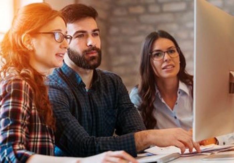 A brown-haired, bearded man sits at a desk with a red-haired woman wearing a plaid shirt and glasses to his right and a brown-haired woman wearing a white shirt and glasses to his left. All three look at a white computer screen and the man has a keyboard in front of him.