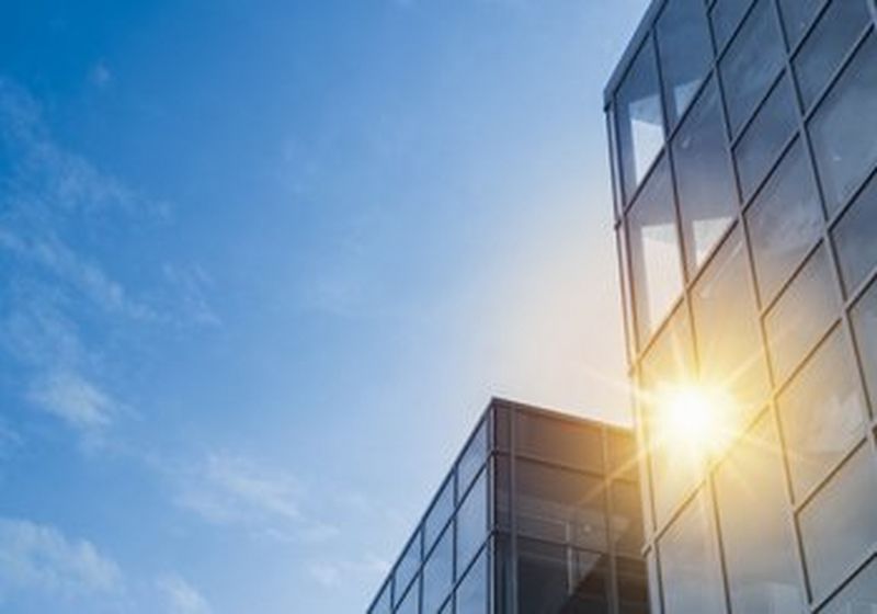 A glass office building on the left, against a blue sky with light clouds. The sun reflects brightly off the building.