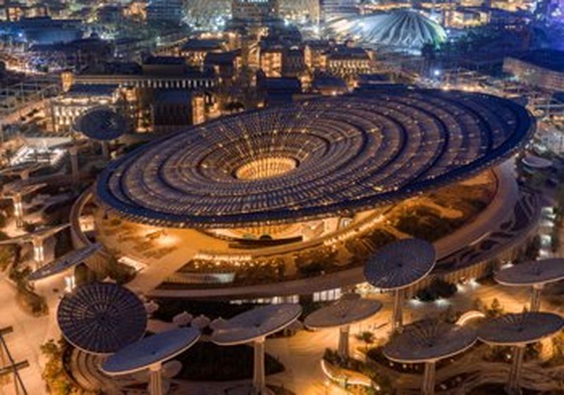 An ariel view of a huge, panelled circular building, emitting golden light that has a cylindrical opening at the top. It is surrounded by smaller versions of itself, sat atop tall metal ‘stems’, all slightly angled in different directions. Beyond the building are other less unusual buildings, lit with white lights.