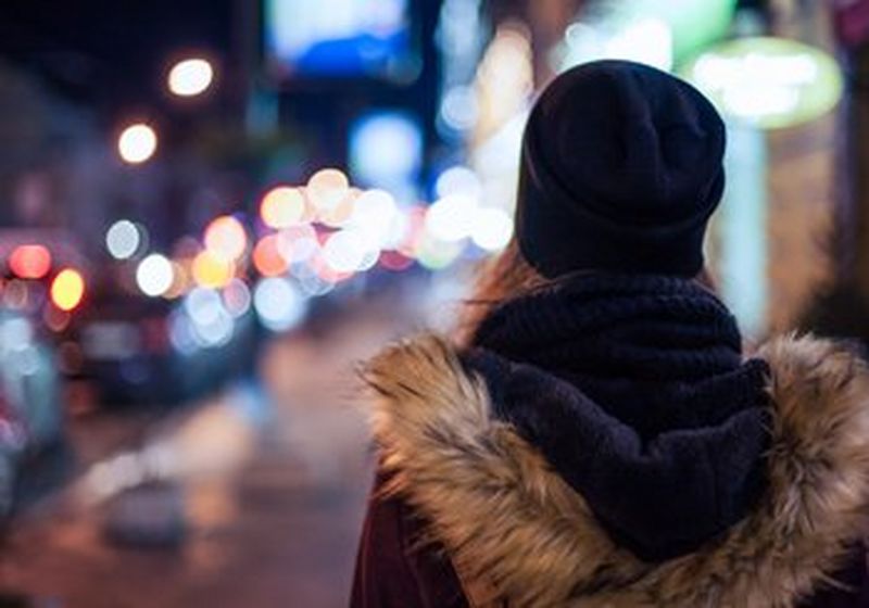 A long-haired person in fur-trimmed parka and navy-blue hat and scarf stands to the right of the picture, facing blurred evening lights, turned away from the camera.