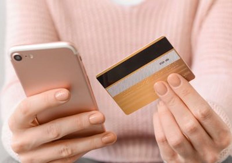 An unseen person in a pale pink sweater holds a pink smartphone in one hand and a credit card in the other.