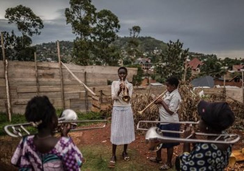 Four girls stand in a circle, playing trombones and trumpets in a dirt yard, in front of a fence.