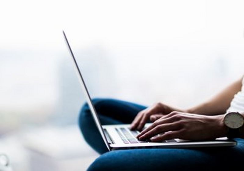 A person in a white t-shirt and blue jeans sits cross-legged, typing on a laptop. Only legs, arms and torso are visible.