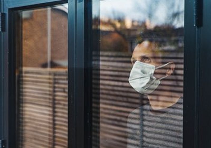 A man wearing a non-surgical face covering and a grey t-shirt, looks out of the window, towards the sky. The glass reflects the outside world, other houses, trees, a fence and the sky.