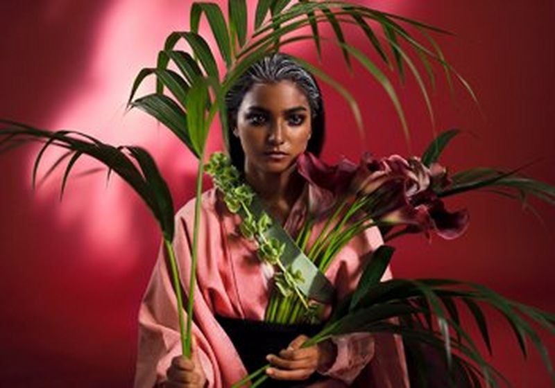 A woman holding green and wine-coloured flowers against a deep pink/purple background and surrounded by green fern-like leaves.