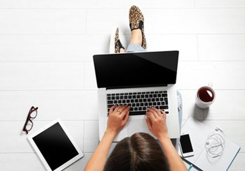 A person viewed from above sits on a white tiled floor with a laptop on their lap. Beside them is a pair of glasses, a tablet, a phone, a notepad, some headphones and a cup of coffee. All white.