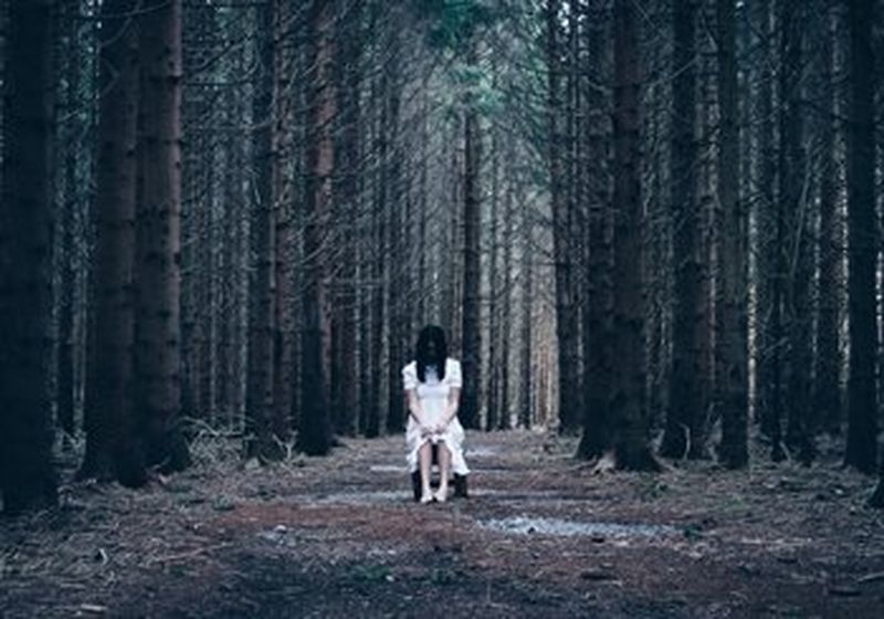 A barefoot woman in a long white dress with long dark hair sits on a wooden stool in a forest, surrounded by tall, bare trees. She has her hands in her lap and is looking down, so her hair is covering most of her face.