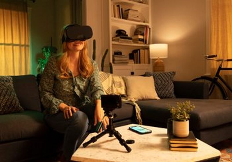 A woman with long blonde hair sits on a couch. She wears a patterned green blouse and grey trousers, and a VR headset. In front of her is a coffee table, upon which is a camera on a tripod, a smartphone, some books and a small plant. Behind her is a white bookcase, two curtained windows and a bicycle.