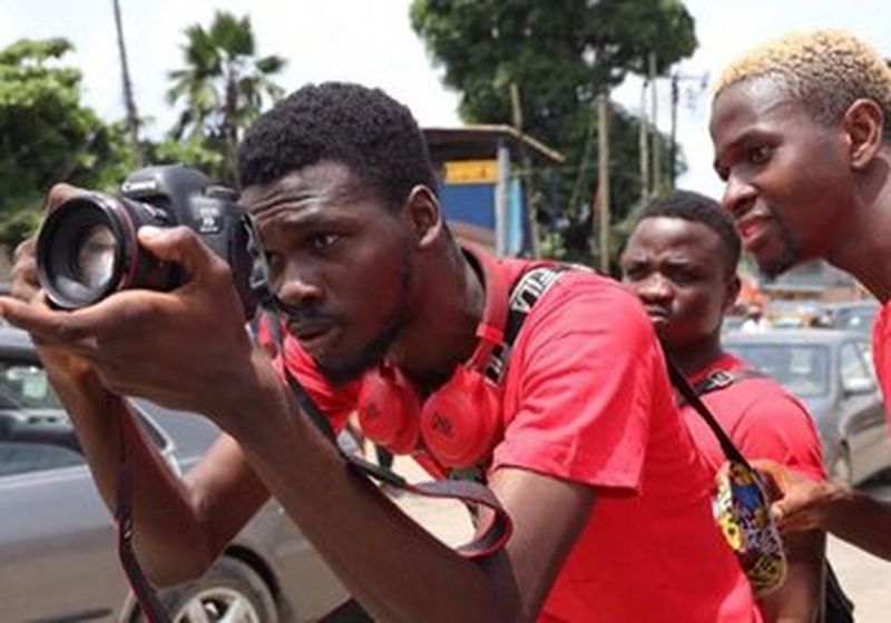 Four young men, one holding a camera, look carefully at the shot they are about to take.