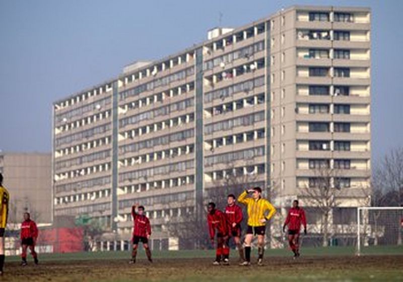 A Sunday league football match being plated on an amateur muddy pitch by players in red or yellow shirts. In the immediate background looms a huge, concrete block of flats.