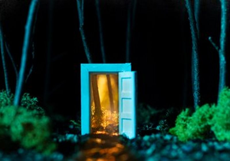 A tiny blue door and doorframe sits in the middle of what appears to be a woodland floor at night. The door is wide open. Revealing more trees, only these trees are illuminated in a golden light.