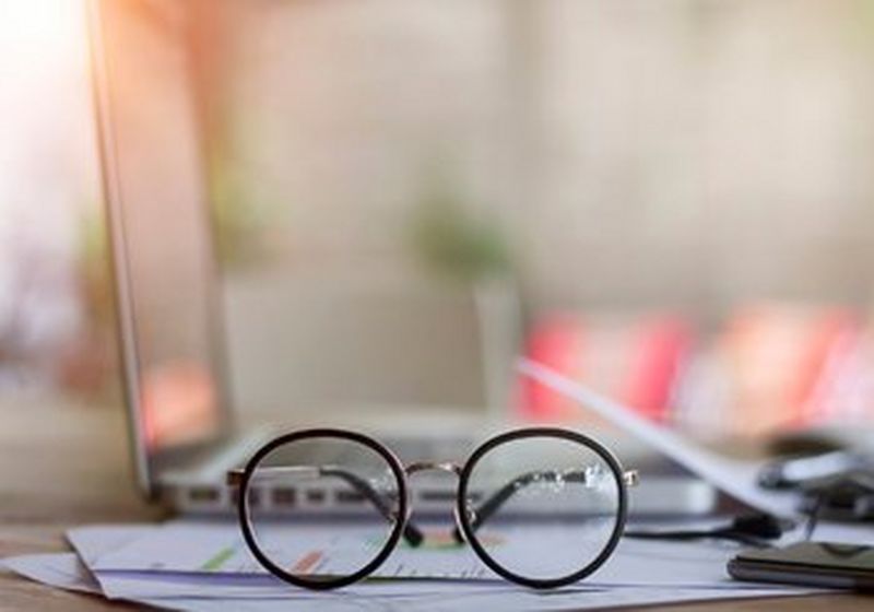 A pair of spectacles sat on top of paperwork, in front of a laptop and next to a phone