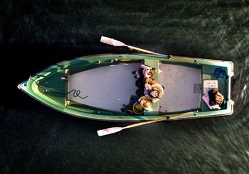 An aerial view of a green rowing boat on the sea, with the front of the boat pointing left and the rear to the right. Oars are sticking out from the top and bottom of the scene. There are three people seated in the centre of the boat and they are looking up to the sky, and the camera.  