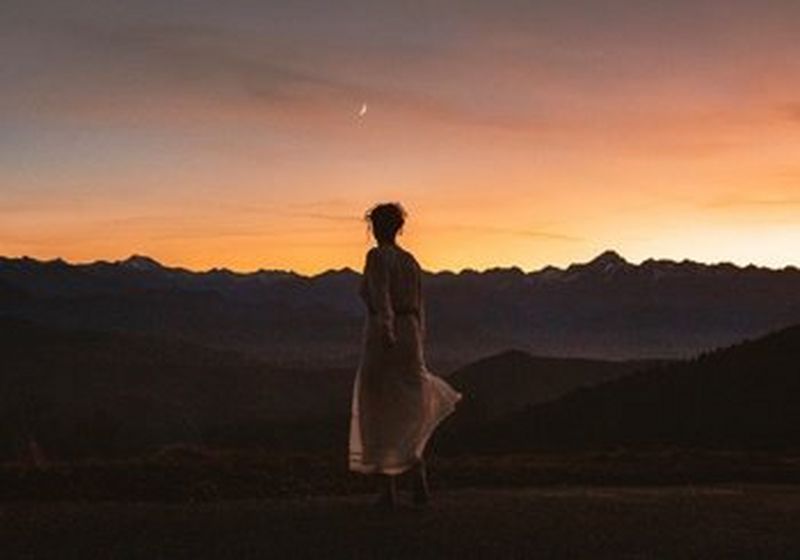 A woman stands with her back to the camera, watching an orange sunset over dark mountains. She wears a pale dress, and her hair is worn up, with tendrils outlined in shadow.