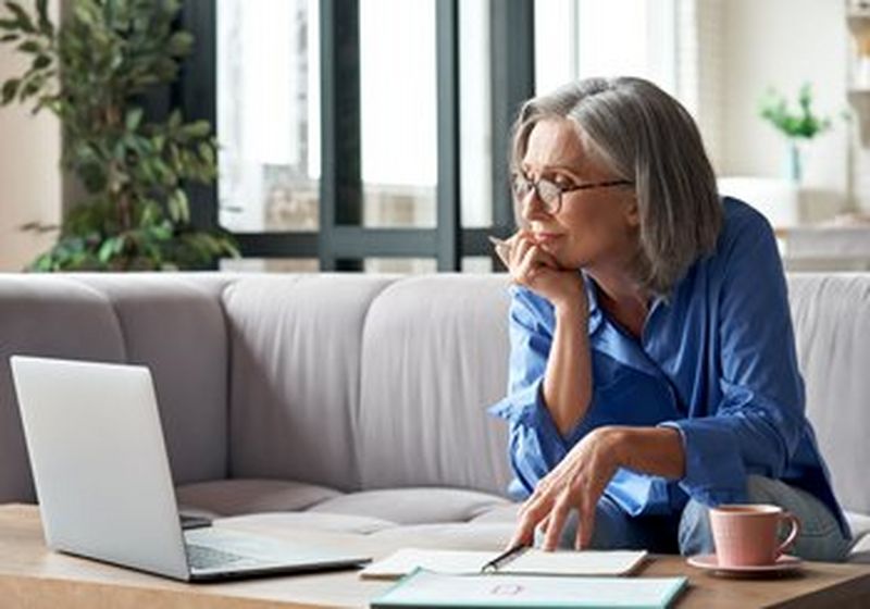 A grey-haired woman in tortoiseshell glasses, seated on a couch, leans forward with her chin resting on her hand to look at her laptop. She has a pink cup and saucer next to her and is wearing a blue shirt.