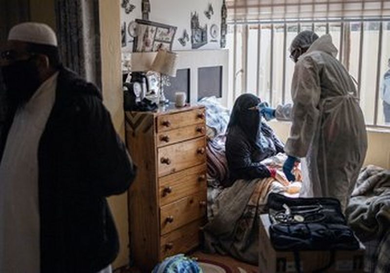 A man dressed in white PPE takes the temperature of a women as she sits up in bed. She is wearing a black niqab and on one side of her is a high brown wood chest of draws with six drawers and a lamp and assorted objects on top. On the other side is a barred window, through whIch you can see the figure of a man wearing a face covering. On the far left of the photo is the side view of a man wearing a white kameez and white topi. He also wears a black protective face covering.