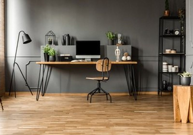 A stylish grey-walled home office, with a light wood desk and chair, several plants, a lamp, objects on black shelves and a desktop computer.