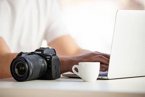 A Canon EOS R6 camera on a desk next to a man typing on a laptop.