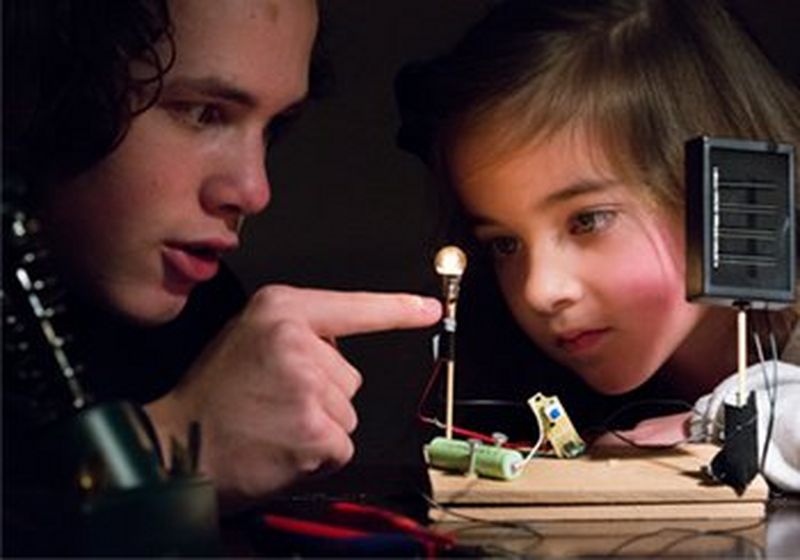 A boy points at a lightbulb, as a small girl watches closely