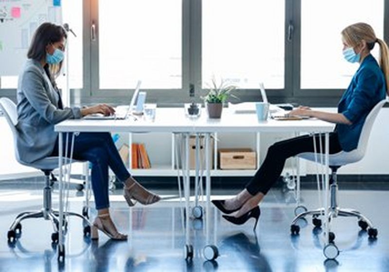 Two women sit opposite each other at a white table, working on their laptops. They are wearing smart, casual clothing and blue disposable, non-surgical face coverings.