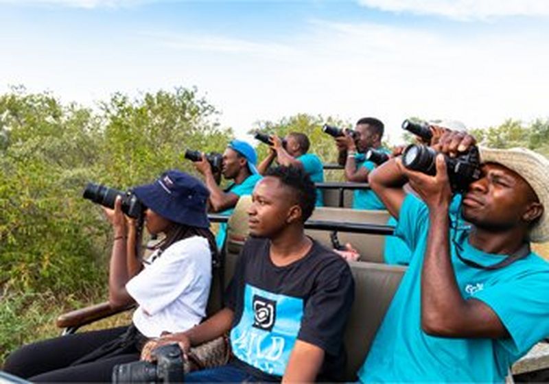 Seven people sit in an open-topped vehicle. Five wear matching bright blue t-shirts and two people at the front of the vehicle do not. The person on the left wears black trousers, a white shirt and a blue bucket hat. Next to them is a man in a black t-shirt with a bright blue ‘wild shots’ logo on the front. He is the only person not holding up a camera as though to take a shot.