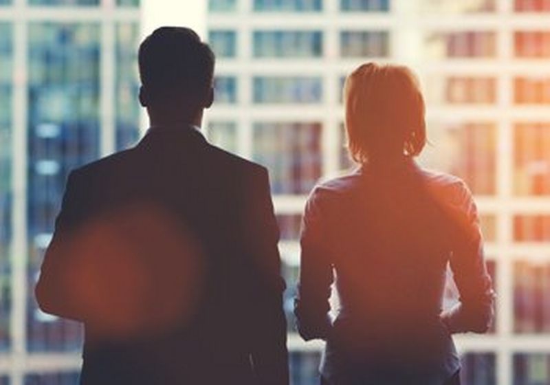 A man and woman, dressed in office attire and standing with their backs to the camera, looking out of a window at other office buildings.