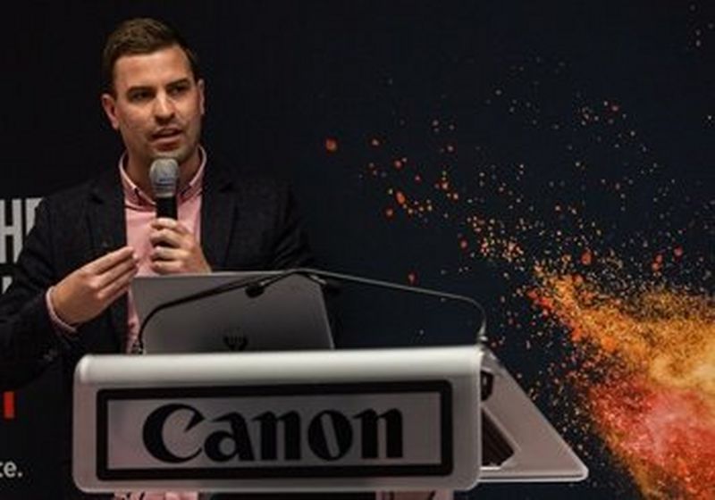 Sam Tatam, giving his keynote speech at a Canon launch event, holding a microphone and standing behind a Canon branded lectern