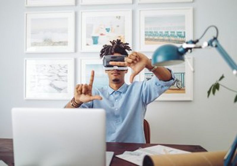 A young man sits at a desk wearing a VR headset and holding his fingers in a ‘frame’ pose. In front of him is a laptop, notebook and lamp. Behind him are nine equally sized pieces of artwork in white frames.