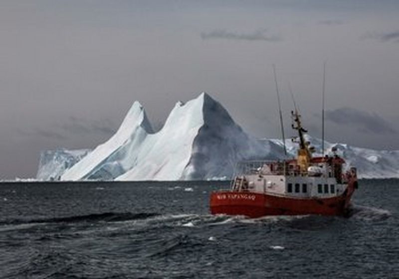 A red boat sails on cold grey water, with a huge floating iceberg behind it.