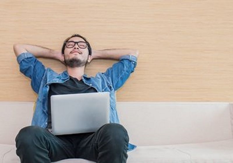 A young man in glasses reclines on a couch with his laptop resting on his lap.