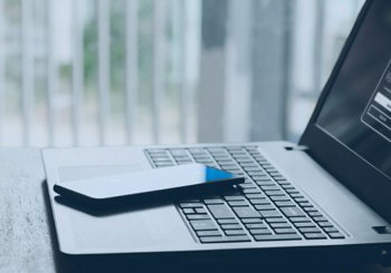 A laptop, viewed from the side, sits open on a table by a window with office-style blinds. Upon its keyboard is a smartphone. The screen shows a login page against a black background.