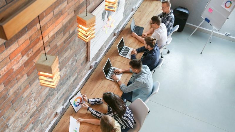 A team of co-workers sit side-by-side at a bench in a modern workspace discussing data on a hardcopy chart. Some are typing on their laptops. A flip chart is positioned behind them