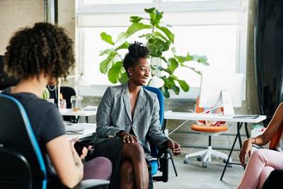 Woman sits on blue chair in front of a plant and talks to two people