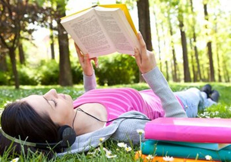 A young woman wearing headphones lies on the grass in a park, reading a book.