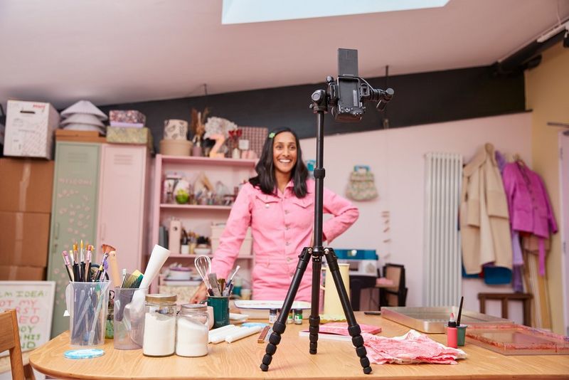 A Canon EOS R50 on a tripod positioned on top of a table to record a woman in a pink jumpsuit surrounded by crafting tools.