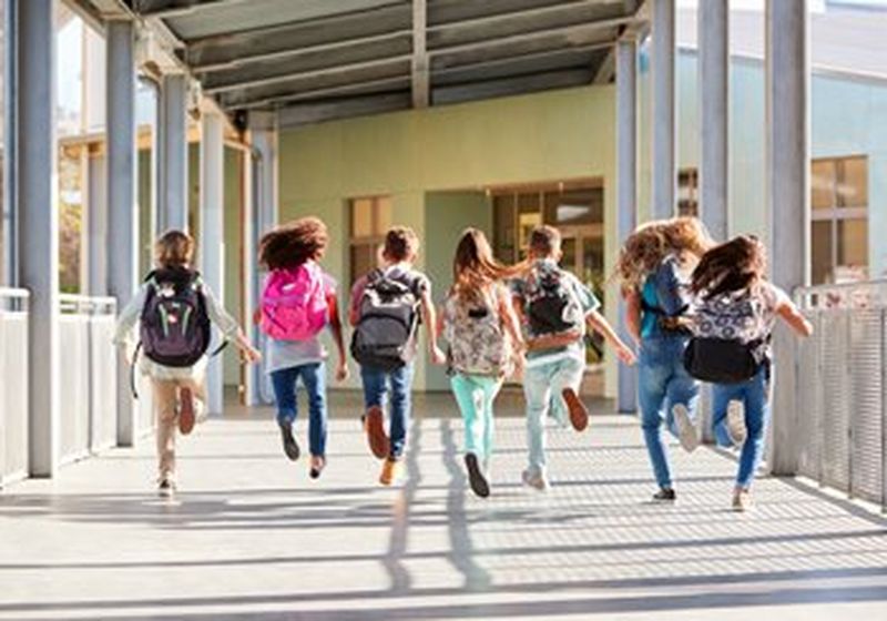 Seven children run side by side down an open corridor towards school doors. Their feet are either one or both off the ground and they all wear a different kind of backpack on their backs. The light suggests the sun is shining. 